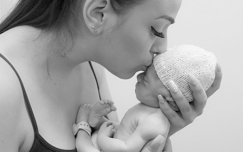 Youg woman holding a tiny baby in a nappy and white hat to her face for a kiss