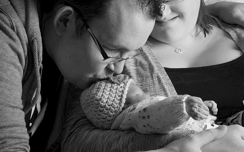 Close up a father kissing the tiny head of his baby with an out of focus mum smiling in the background