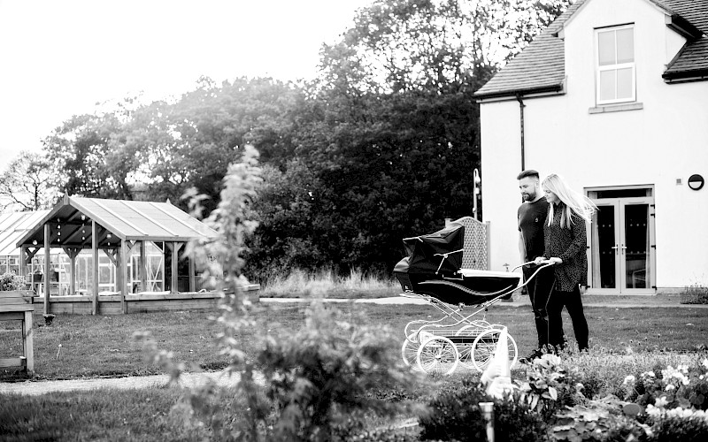 Long shot of a couple pushing a traditonal coach built pram in the gardens of a white house