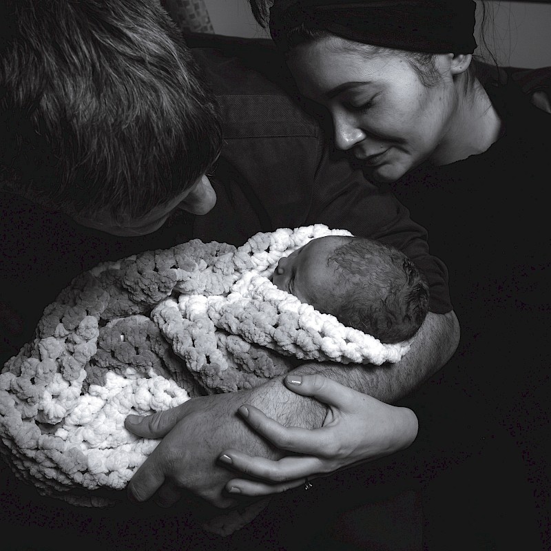 Dark monochrome image of baby being cradled by parents and wrapped in a shawl